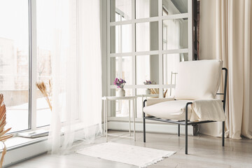 A white faux leather armchair near the window next to a small table with flowers. Part of the interior with copy space.High quality photo