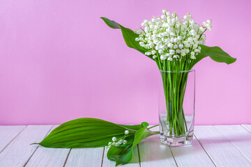 A bouquet of white lily of the valley flowers in a glass on a pink background