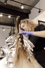 Back view of woman sitting in beauty salon with hair wrapped in foil. Hairdresser colorist looking at head of client.