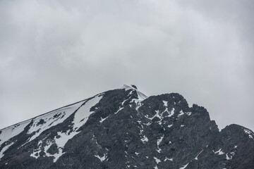 Monochrome scenery with high mountain peak with snow cornice in gray cloudy sky at overcast. Dark atmospheric mountain landscape with large pinnacle in lead gray sky at rainy weather in grayscale.