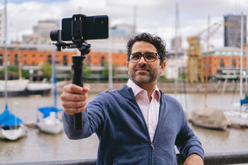 Middle-aged Latin man with glasses documenting with his cell phone a river port on a levee in the middle of the city.