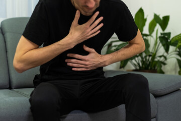 The man is sitting on a gray couch and holding his belly. Medicine and health concept, stomach problems. The man suffers from stomach ache, gastric problems. Abdominal pain, suffering and pain