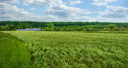 Fototapeta na wymiar Panoramic view of green wheat field and solar panels under beautiful sky with clouds.