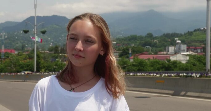 Portrait of cute blonde teen girl tourist standing on highway with mountain background. Teenager smiling enjoying road trip having rest stop in amazing place. Travel tourism vacation car trip concept.