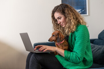 One blond woman sitting on the couch in the living room working on the laptop with her Dachshund dog 