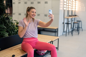 Fototapeta na wymiar Happy satisfied athletic young woman sitting on bench in gym's dressing room using smart mobile phone, waving on video call to say hi before going to fitness class workout, looking satisfied and happy