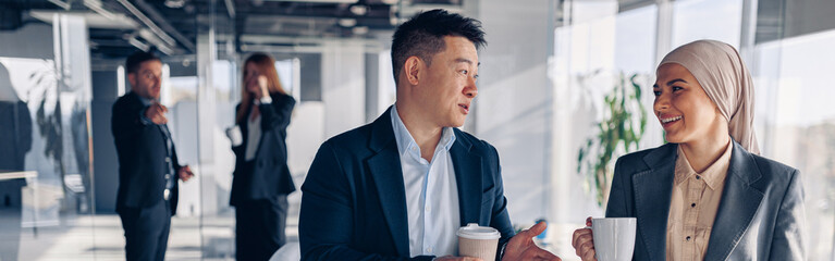 Two happy multiracial colleagues talking and drinking coffee during break time in office