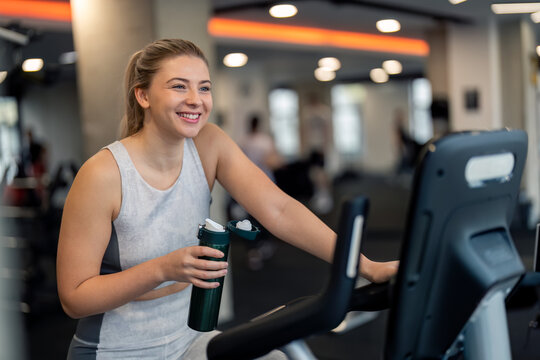 Young confident fit woman gym member on stationary exercise bike machine taking a break from cycling exercising holding water bottle staying hydrated and positive during workout at gym.