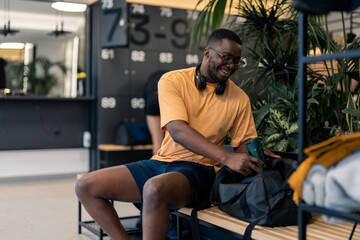 Fototapeta na wymiar Handsome athletic man with headphones looking satisfied sitting on bench in locker room at gym, packing water bottle in sports gym bag after finished workout session, preparing to go home.