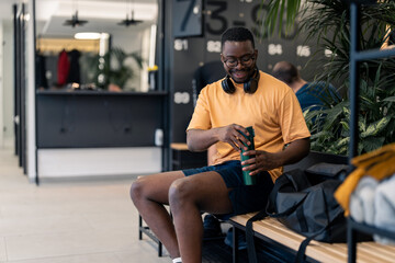 Fototapeta na wymiar Young athlete man sportsperson in yellow t-shirt with eyeglasses and headphones holding opening water bottle sitting on bench in locker room at gym with smile after completed workout training session.