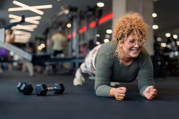 Determined smiling young woman gym fitness client in plank position doing exercises for strong core abdominal muscles having dumbbell weight set next to her focused on fitness goal at gym.