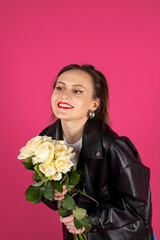 Cool biker girl wearing a leather jacket and jeans posing with a bouquet of white roses. Isolated on pink background.