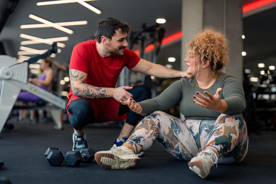 Confident smiling male gym coach professional fitness trainer supporting his client active plus-size young woman discussing future exercising individual training plan with her in gym fitness center.