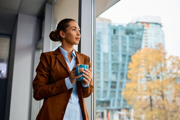 Young businesswoman daydreaming while having cup of coffee by window in her office.