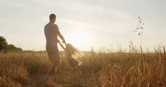 Happy young father have fun with his little daughter spending time together in a wheat field at sunset. Dad spinning his daughter holding hands laughing during golden hour.