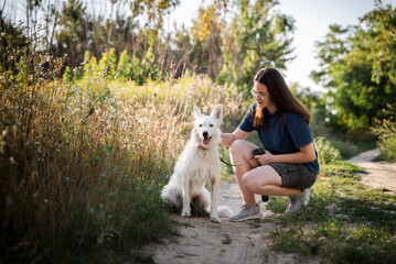 A girl in glasses is stroking a white dog on a dirt road in the forest.