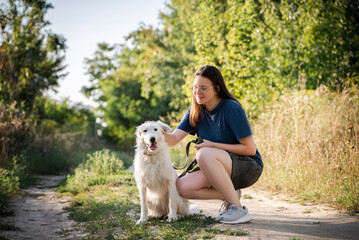 The girl is stroking a white dog against the background of the forest. They are on a dirt road.