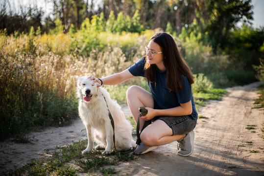 A woman strokes a white dog against the background of the forest. The dog is sitting on a dirt road.