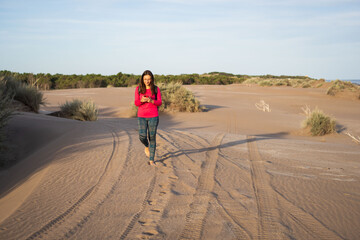 Obraz premium Young bespectacled Latina woman walking face forward using her cell phone on the dunes of a South American beach in Buenos Aires province.