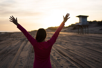 Fototapeta na wymiar Backlit woman raising her arms at sunset against the light on a beach with lifeguard hut