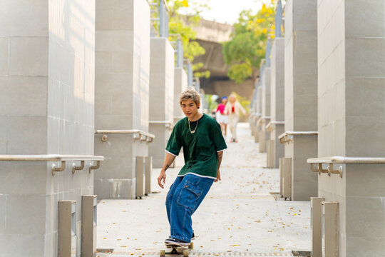 Group of Young Asian man and woman friends skating on longboard skate at public park on summer vacation. Stylish people have fun urban lifestyle practicing outdoor sport skateboarding at city street.