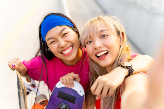 Asian woman friends using mobile phone taking selfie together during skating on longboard skate in the city. Stylish girl have fun outdoor lifestyle skateboarding on city street on summer vacation.