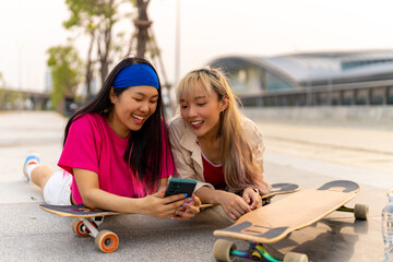 Asian woman friends mobile phone with internet together during skating on longboard skate on summer vacation. Stylish girl enjoy urban outdoor lifestyle extreme sports and portable device in the city.