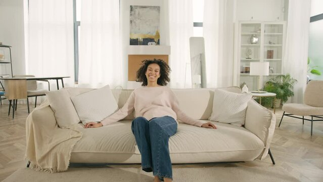 Afro girl laughing, enjoying rest on cozy sofa, feeling overjoyed, celebrating move in day to home, rented flat. Design interior furniture. Happy African American woman jumping on modern soft couch 4K