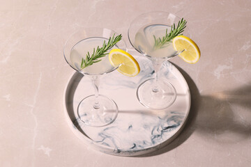 Tray with martini glasses of fresh cocktail, rosemary and lemon slices on beige marble table