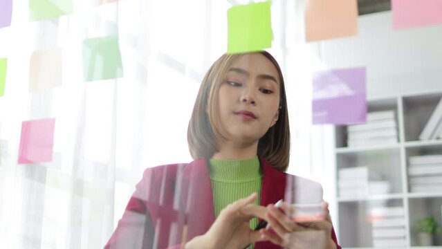 Asian businesswoman scheduling work strategy, planning work in the office with paper stickers on the glass.