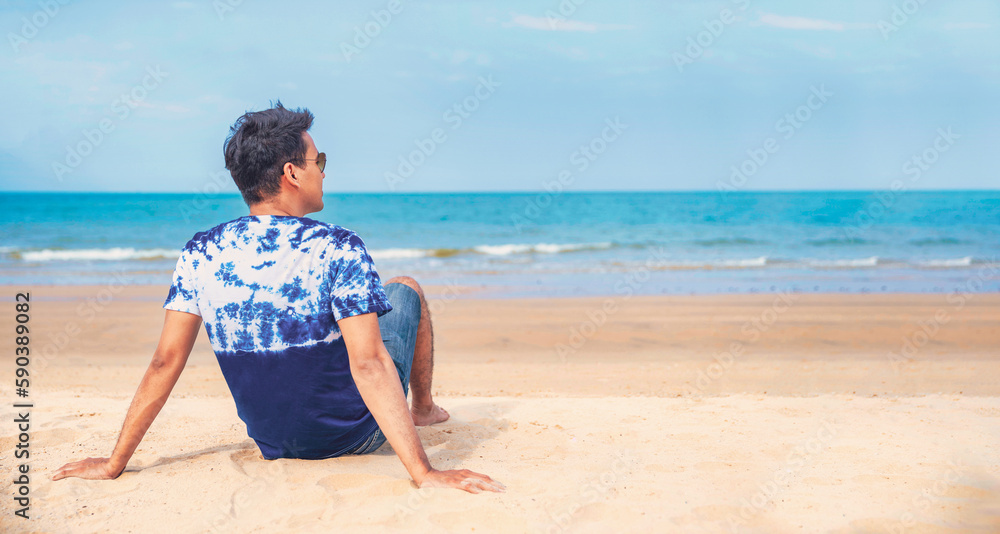 Wall mural Happy young man in casual clothing wearing sunglasses and smiling at the beach. Handsome guy laughing at the beach and looking away. Cheerful guy enjoying summer vacation 
