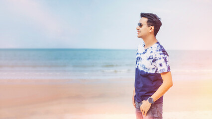 Happy young man in casual clothing wearing sunglasses and smiling at the beach. Handsome guy laughing at the beach and looking away. Cheerful guy enjoying summer vacation 
