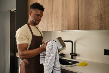 Young african man drying stainless steel pot with kitchen towel in a kitchen.