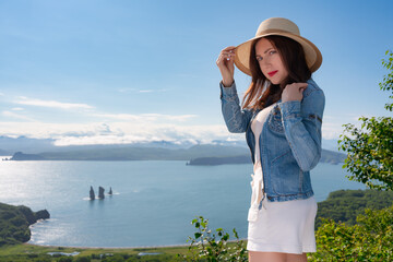 Woman in straw hat, denim jacket and white shorts standing on against backdrop of sea on sunny day