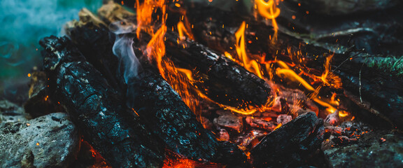 Vivid smoldered firewoods burned in fire close-up. Atmospheric warm background with orange flame of campfire and blue smoke. Full frame image of bonfire. Beautiful whirlwind of embers and ashes in air