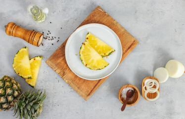 Closeup shot of two pieces sliced yellow pineapples on white dish on wood cutting board placed on table with other ingredients aroma oil in glass vial, pepper bottle, salt in wooden bowl and onion