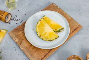 Closeup shot of two pieces sliced yellow pineapples on white dish on wood cutting board placed on table with other ingredients aroma oil in glass vial, pepper bottle, salt in wooden bowl and onion
