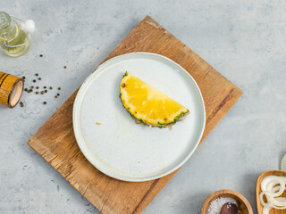 Closeup shot of two pieces sliced yellow pineapples on white dish on wood cutting board placed on table with other ingredients aroma oil in glass vial, pepper bottle, salt in wooden bowl and onion