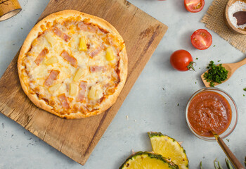 Top view shot of delicious tasty juicy of Hawaiian traditional Italian crust thin crispy ham and pineapple pizza placed on wooden cutting board with ingredients sliced tomatoes, ketchup and salt