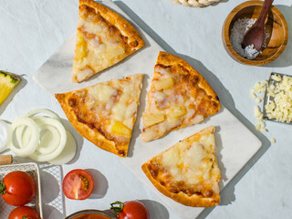 Top view shot sliced triangle pieces of delicious tasty homemade Hawaiian traditional Italian crust thin crispy ham and pineapple pizza placed on dish with ingredients tomatoes cheese salt on table