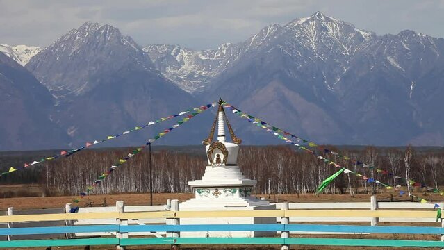 Buddhist Stupa decorated with multi-colored prayer flags in the Tunka foothill valley against the backdrop of the Eastern Sayan Mountain ranges on a sunny spring day. Buryatia