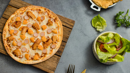 Top view shot of delicious tasty juicy sliced sausage cheesy pizza placed on wooden board around with other fast food crispy fried chicken, seasoning ingredients and fresh raw organic vegetables