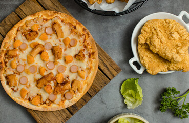 Top view shot of delicious tasty juicy sliced sausage cheesy pizza placed on wooden board around with other fast food crispy fried chicken, seasoning ingredients and fresh raw organic vegetables