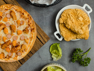 Top view shot of delicious tasty juicy sliced sausage cheesy pizza placed on wooden board around with other fast food crispy fried chicken, seasoning ingredients and fresh raw organic vegetables