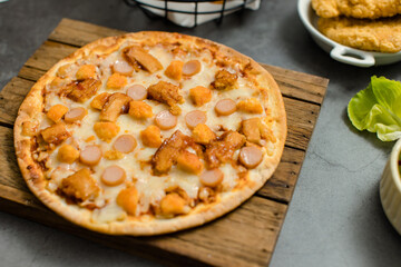 Top view shot of delicious tasty juicy sliced sausage cheesy pizza placed on wooden board around with other fast food crispy fried chicken, seasoning ingredients and fresh raw organic vegetables
