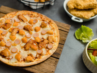 Top view shot of delicious tasty juicy sliced sausage cheesy pizza placed on wooden board around with other fast food crispy fried chicken, seasoning ingredients and fresh raw organic vegetables
