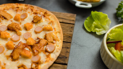 Top view shot of delicious tasty juicy sliced sausage cheesy pizza placed on wooden board around with other fast food crispy fried chicken, seasoning ingredients and fresh raw organic vegetables