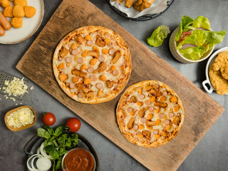 Top view shot of two delicious tasty juicy sliced sausage cheesy pizza placed on wooden board around with other fast food crispy fried chicken, seasoning ingredients and fresh raw organic vegetables