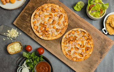 Top view shot of two delicious tasty juicy sliced sausage cheesy pizza placed on wooden board around with other fast food crispy fried chicken, seasoning ingredients and fresh raw organic vegetables