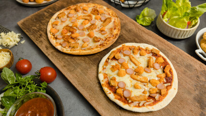 Top view shot of two delicious tasty juicy sliced sausage cheesy pizza placed on wooden board around with other fast food crispy fried chicken, seasoning ingredients and fresh raw organic vegetables